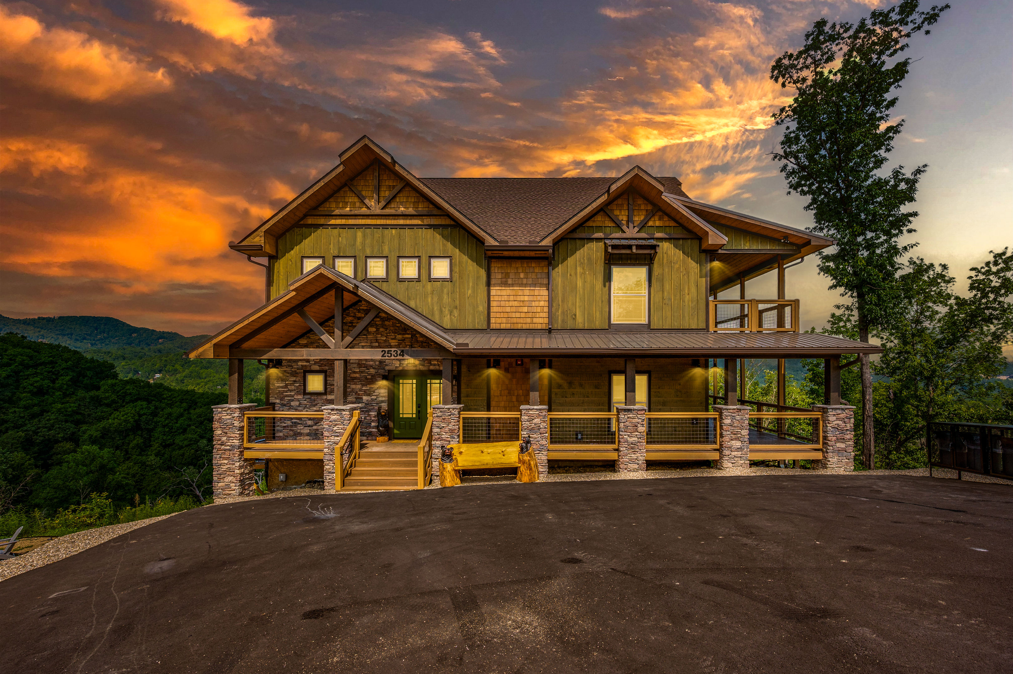 Dramatic sunset over a modern mountain cabin with green wood siding, stone pillars and a wraparound porch overlooking forested hills