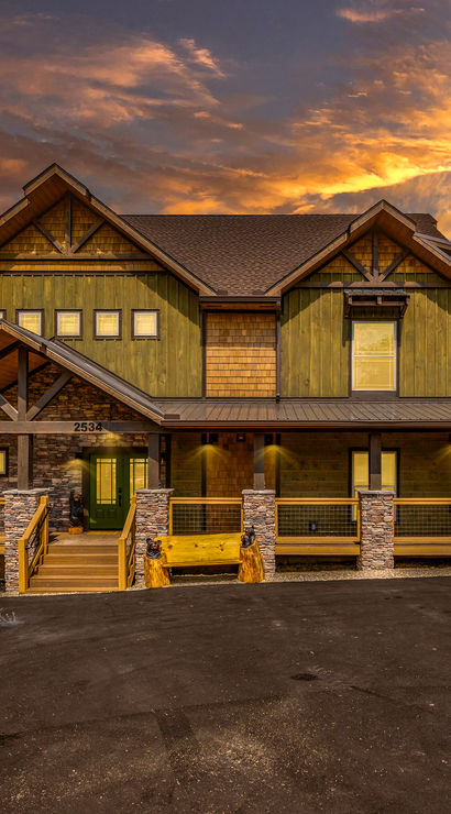 Dramatic sunset over a modern mountain cabin with green wood siding, stone pillars and a wraparound porch overlooking forested hills