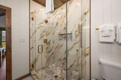 Spa-like modern bathroom with glass-enclosed shower, marble-look wall tiles, gold hardware, hexagon pebble floor, built-in niche and folded white towels on board-and-batten walls.