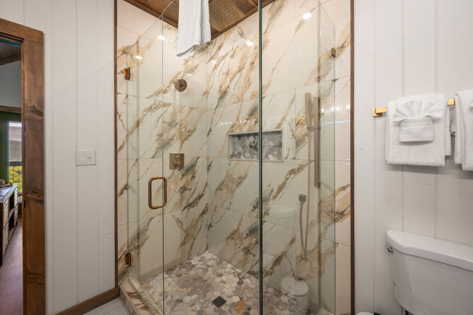 Spa-like modern bathroom with glass-enclosed shower, marble-look wall tiles, gold hardware, hexagon pebble floor, built-in niche and folded white towels on board-and-batten walls.