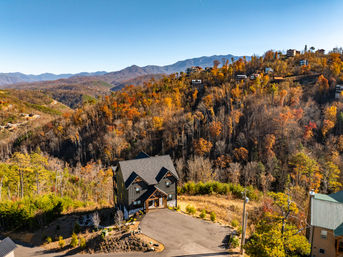 Aerial view of a modern mountain house perched on a hilltop, surrounded by vibrant fall foliage, a winding driveway and distant blue mountain ridges under a clear sky