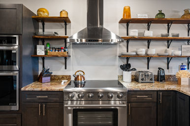 Contemporary kitchen with stainless steel range and hood centered between dark wood cabinets, granite countertops, open wooden shelves stocked with white dishes, spices and decorative jars; copper kettle, toaster and utensil jar on the counter.