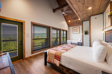 Cozy rustic mountain-view cabin bedroom with vaulted wood ceiling, large windows and balcony, queen bed with white linens and a patchwork throw, wooden floors and modern farmhouse decor.