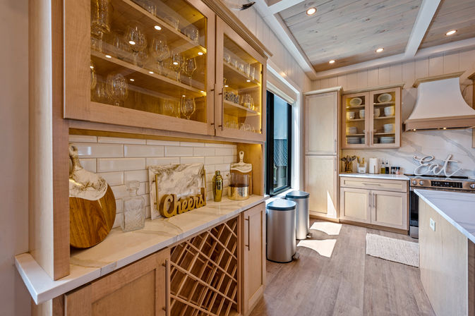 Sunlit modern farmhouse kitchen with light oak cabinets, glass-front cupboards of stemware, marble countertops, built-in wine rack, stainless bins, and decorative “Cheers” and “Eat” signs.