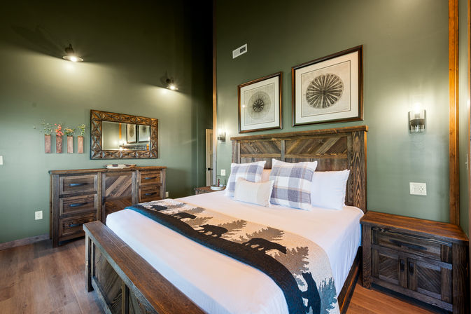 Cozy rustic bedroom with king bed featuring a dark wood headboard, plaid pillows and a wildlife‑silhouette throw, green painted walls, matching wooden dresser and nightstands, framed prints and wall sconces.