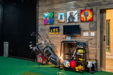 Indoor sports practice room with green turf and wood-paneled wall, two golf bags with clubs, a monitor and keyboard on a small desk, shelving of soccer/volleyballs and equipment, and colorful pop-art canvases above.