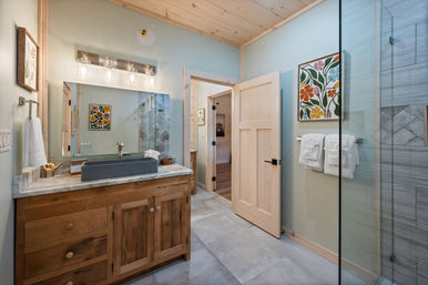 Bright, airy rustic bathroom with light blue walls, natural wood vanity and marble countertop, rectangular vessel sink, large mirror and industrial-style vanity lights, glass-enclosed tiled shower, floral wall art and neatly folded white towels.