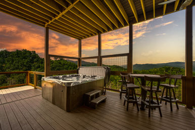 Covered wooden deck with bubbling hot tub and bar-height table, panoramic forested mountain view at sunset