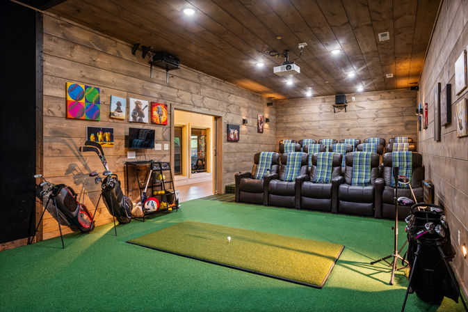 Wood-paneled indoor golf simulator room with green turf hitting mat and practice tee, ceiling projector, tiered leather recliners draped with plaid throws, golf bags, equipment racks and colorful wall art