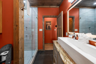Rustic mountain-style bathroom with bold red walls, glass walk-in shower, reclaimed wood trim and barn door, long white rectangular sink on a granite countertop, illuminated mirror and neatly folded white towels.