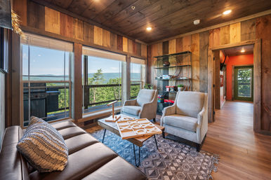 Cozy rustic mountain cabin living room with wood-paneled walls, leather sofa, two beige armchairs, patterned rug and coffee table with tic‑tac‑toe, large windows framing a panoramic green mountain view and balcony grill.