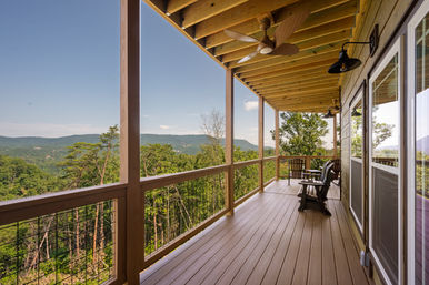 Covered wooden porch with Adirondack chairs and ceiling fans overlooking a forested valley and distant blue mountain ridges under a clear sky.