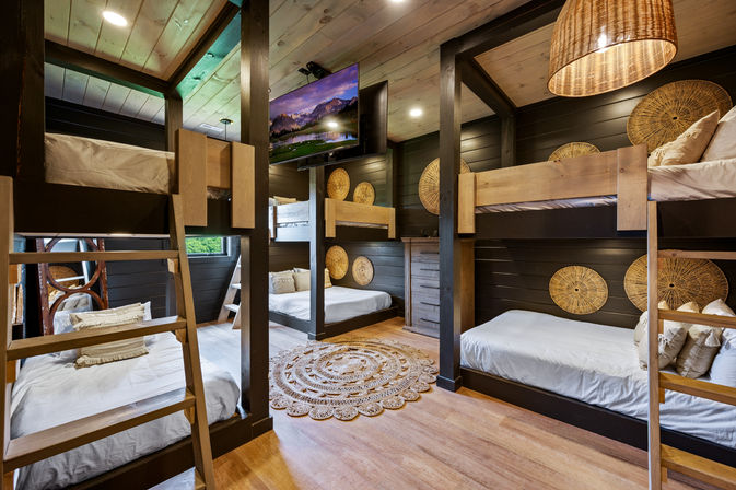 Cozy rustic cabin bunk room with multiple wooden bunk beds, woven rattan wall accents, round braided rug, pendant wicker light, hardwood floors and wall-mounted TV — ideal vacation-lodge sleeping space.