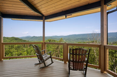 Inviting rocking chairs on a covered wooden cabin deck overlooking a lush forested valley and distant blue mountain ridges under a clear sky.
