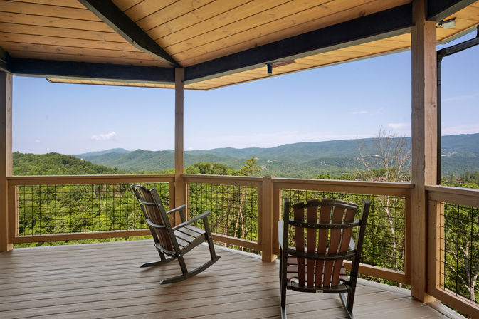 Inviting rocking chairs on a covered wooden cabin deck overlooking a lush forested valley and distant blue mountain ridges under a clear sky.
