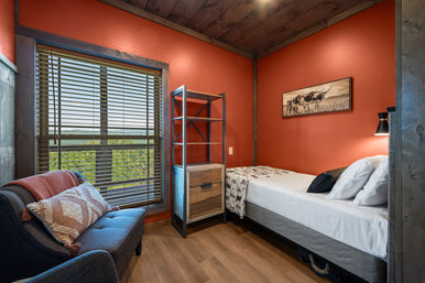 Cozy rustic guest bedroom with warm orange walls and wood ceiling, single bed with white linens and patterned throw, metal‑wood shelving, navy armchair, and a large window with blinds overlooking green treeline.