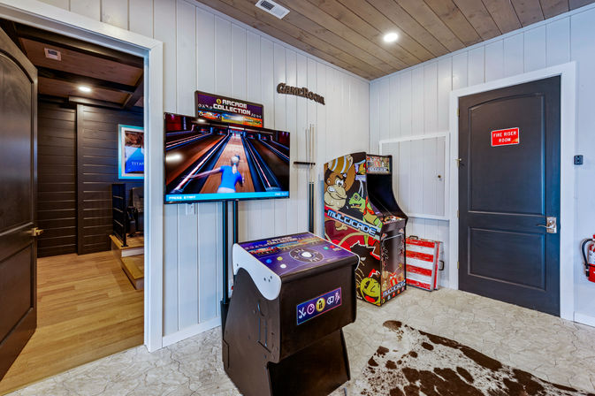 Playful indoor game room with wall-mounted TV showing a virtual bowling game, a cocktail-style arcade cabinet, colorful retro arcade machine, wood-paneled ceiling and cowhide rug.