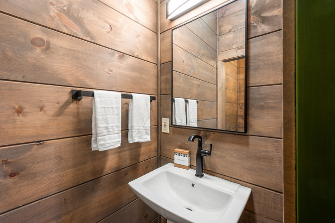 Cozy rustic cabin-style bathroom with horizontal wood-plank walls, white wall-mounted sink, matte black faucet and rectangular mirror, and a black towel bar holding two folded white towels.