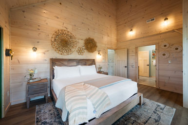 Cozy light-wood cabin bedroom with vaulted pine walls, king bed with crisp white linens and striped throw, woven wall decor above the headboard, wooden nightstands, warm wall sconces and a patterned area rug.