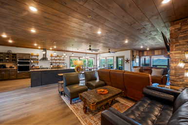 Cozy open-plan rustic mountain living room and kitchen with wood-plank ceiling, leather sofas, stone fireplace, island with pendant lights, and panoramic mountain views.