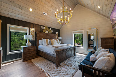 Cozy luxury rustic bedroom in a mountain cabin with vaulted shiplap ceiling, wooden king bed, gold chandelier, bedside tables, plush rug and windows framing lush forest views.