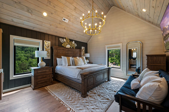 Cozy luxury rustic bedroom in a mountain cabin with vaulted shiplap ceiling, wooden king bed, gold chandelier, bedside tables, plush rug and windows framing lush forest views.