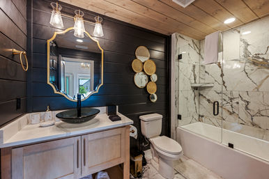 Modern farmhouse bathroom with black shiplap wall, gold-trim mirror, black vessel sink on a light-wood vanity, woven round wall decor, and marble-tiled shower with glass door and white tub.