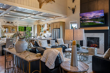 Cozy open-concept modern farmhouse living room and kitchen with leather sofas, chunky knit throw, oversized vase of branches, wood-beamed ceiling, fireplace and wall-mounted TV showing a mountain scene.