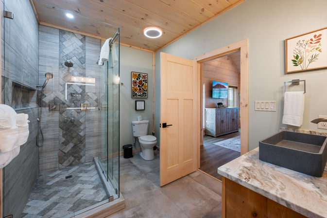 Cozy modern rustic cabin-style bathroom with glass-enclosed herringbone-tile shower and dual showerheads, wood-paneled ceiling, marble countertop with rectangular stone sink, toilet, towel rack, and open door to a wood-finished bedroom