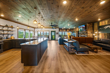 Open-concept mountain lodge interior with a large marble-top kitchen island, hanging pendant lights, wood-plank ceiling and floors, dining area and cozy living room featuring a stone fireplace, leather seating, and panoramic windows overlooking green hills.