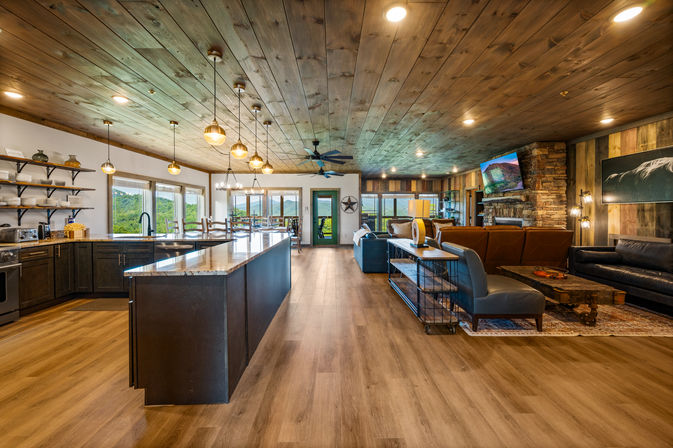 Open-concept mountain lodge interior with a large marble-top kitchen island, hanging pendant lights, wood-plank ceiling and floors, dining area and cozy living room featuring a stone fireplace, leather seating, and panoramic windows overlooking green hills.