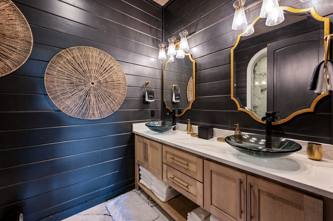 Modern luxury bathroom with dark horizontal shiplap walls, double wood vanity and marble countertop, two glass vessel sinks with black faucets, gold-framed mirrors, glass pendant lights and woven round wall decor.
