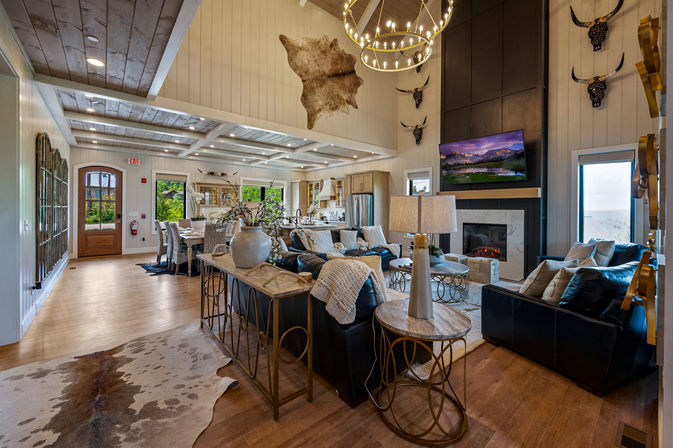 Cozy rustic-chic open-concept living room and kitchen with vaulted wood-beamed ceiling, circular chandelier, leather sofas, cowhide rug, stone fireplace with wall-mounted TV, dining table, decorative antler mounts, and large windows with a scenic water view.