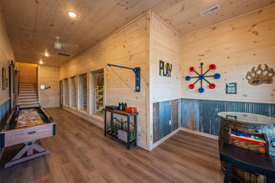 Rustic wood-paneled cabin game room with shuffleboard table, wall-mounted ring toss and decor, barrel game table, corrugated metal wainscoting and staircase