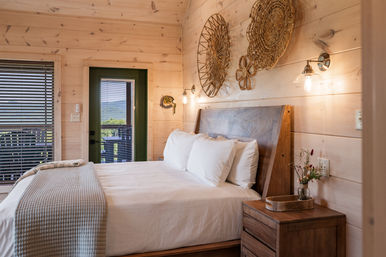 Cozy rustic wood-paneled cabin bedroom with a neatly made queen bed, woven wall art, wooden bedside table and glowing wall sconces; green door opens to a balcony with mountain views.