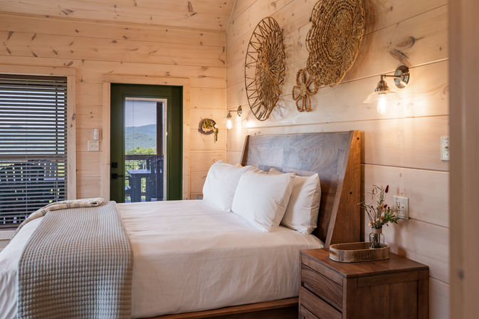 Cozy rustic wood-paneled cabin bedroom with a neatly made queen bed, woven wall art, wooden bedside table and glowing wall sconces; green door opens to a balcony with mountain views.