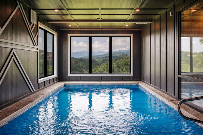 Modern indoor pool with bright blue water and large windows framing rolling green mountains and distant peaks under a blue sky.