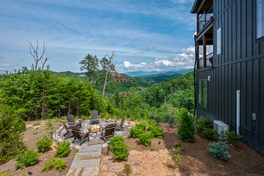 Cozy fire pit with Adirondack chairs on a stone patio beside a modern dark-sided mountain cabin, overlooking a lush forested valley and layered blue mountain ridges under a bright, partly cloudy sky.