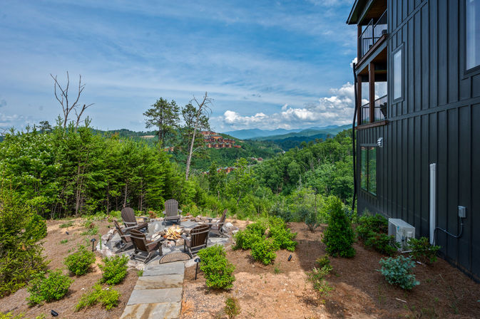 Cozy fire pit with Adirondack chairs on a stone patio beside a modern dark-sided mountain cabin, overlooking a lush forested valley and layered blue mountain ridges under a bright, partly cloudy sky.