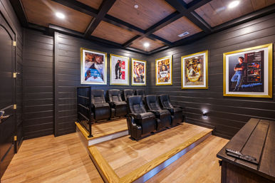 Cozy residential home theater with tiered wooden platforms, six black leather recliners, dark shiplap walls, framed movie posters and recessed ceiling lights.