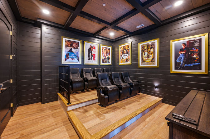 Cozy residential home theater with tiered wooden platforms, six black leather recliners, dark shiplap walls, framed movie posters and recessed ceiling lights.