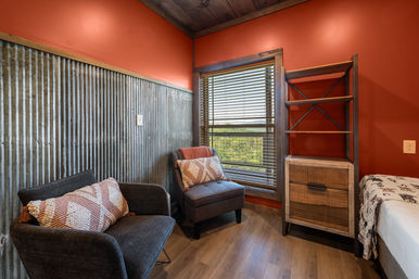 Cozy rustic cabin bedroom nook with red walls and corrugated metal wainscoting, two gray upholstered chairs with patterned pillows by a window with wood blinds overlooking treetops, open wood shelving and a bedside chest.