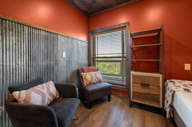 Cozy rustic cabin bedroom nook with red walls and corrugated metal wainscoting, two gray upholstered chairs with patterned pillows by a window with wood blinds overlooking treetops, open wood shelving and a bedside chest.