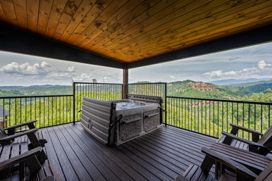 Covered wooden deck with bubbling hot tub and Adirondack chairs overlooking lush forested mountains and distant hillside lodges under a cloudy sky