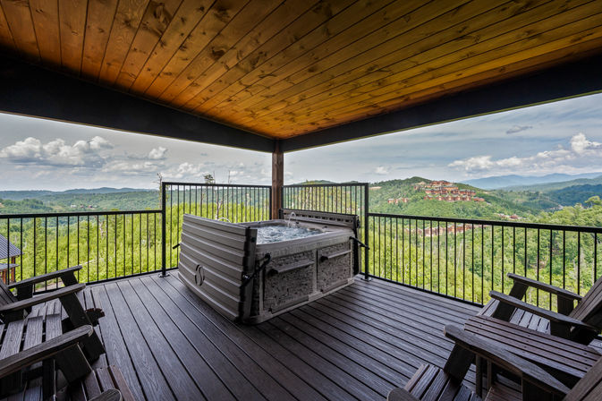 Covered wooden deck with bubbling hot tub and Adirondack chairs overlooking lush forested mountains and distant hillside lodges under a cloudy sky