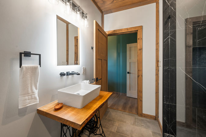 Modern rustic bathroom interior with a white rectangular vessel sink on a live-edge wooden vanity, backlit mirror and matte black wall-mounted faucet, towel on black holder, open wood-trimmed door to teal hallway, gray tile floor and black marble shower panel.