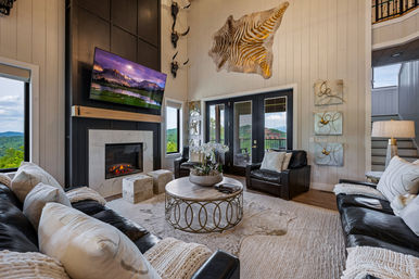 Cozy modern farmhouse living room with vaulted shiplap walls, stone fireplace and wall-mounted TV, leather sofas, round metal coffee table, zebra-hide wall art and glass doors framing a mountain view.