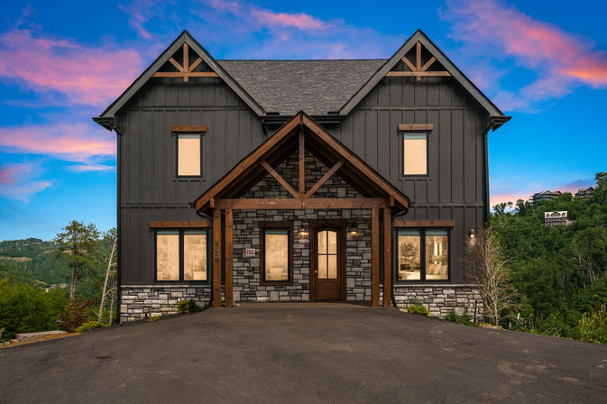Modern two-story mountain home with dark vertical siding, stone lower facade and timber gabled entry, set on a wooded hillside under a pink and blue sunset sky.
