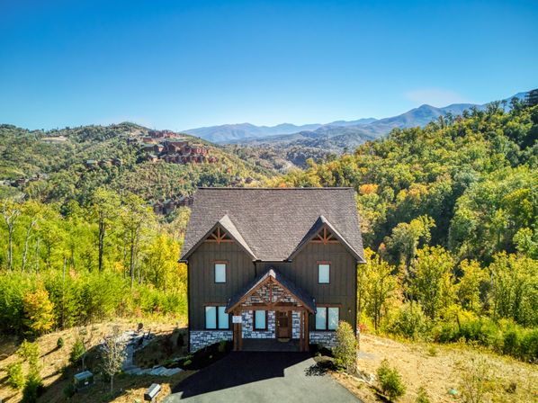 Rustic two-story mountain cabin with stone trim on a sunny hillside, surrounded by green and golden forest and panoramic mountain ridges under a clear blue sky