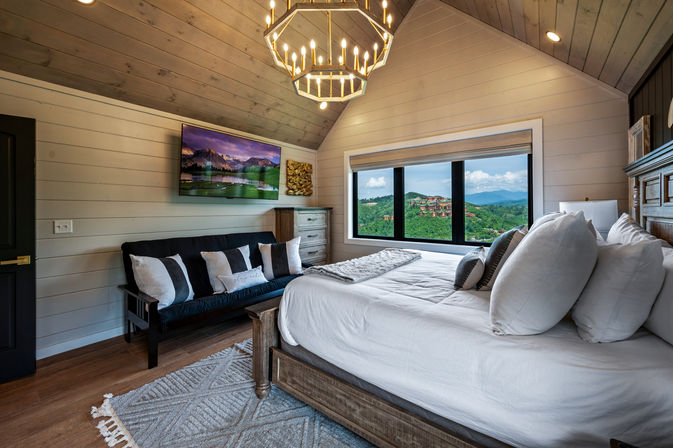 Cozy rustic-modern bedroom with a large bed, geometric chandelier, black sofa with pillows, wall-mounted TV and a wide picture window showcasing lush green hills and distant blue mountains.